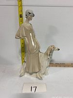 Front view of Royal Doulton figurine with woman in beige dress, gray hat, standing beside a long-haired white and brown dog, measuring around 13 inches tall.