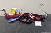 Photo showing two ruby red glass swan dishes and a ceramic boat figurine on a black surface.