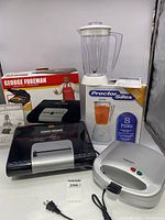 Photo showing George Foreman grill in front with box, Proctor Silex blender box behind and Cuisinart sandwich grill to side