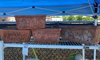 Front view of four terracotta rectangular planters on metal shelving unit under blue canopy, three with leaf designs, one with animal relief