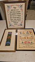All three framed needlepoint and cross stitch artworks arranged together on a surface, featuring the quote piece, floral piece, and sampler piece.