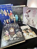 Five antique reference books arranged on a dark surface including titles on Royal Doulton figures, American art pottery, costume history, doll marks, and world antiques.
