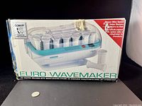 Photo of the front of the box showing the Conair Euro Wavemaker steam rollers set with rollers loaded inside a white and turquoise base with a clear plastic cover.