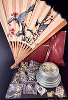 Photo showing leather purse, decorative folding fan, brooches mostly in plastic packaging, and frosted glass bowls.