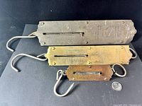 Three vintage metal spring balance scales in different sizes laid flat on black background with a quarter coin for scale. Scales have hooks and embossed text worn from age.
