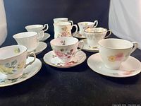 Twelve assorted tea cups and mugs arranged in two rows on a black surface, various floral and gold trimmed designs visible