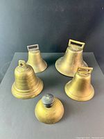 Front view of five vintage brass bells arranged on a black surface, showing varying sizes and shapes including one with a black knob top.