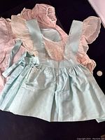 Three vintage handmade doll dresses laid flat: light blue with embroidery and ruffled straps, pink dress with puffed sleeves, and an off-white long dress