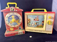 Front view of Fisher-Price Teaching Clock and Giant Screen Music Box with yellow handles, showing clock face and illustrated music box screen.
