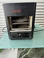 Front view of black Gemelli infrared grill showing the grilling racks inside the opening, control knob and digital display on front panel.