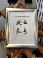 Framed lithograph showing all four monkeys, in silver frame on wooden background.