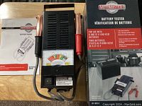 Photo of Motomaster battery tester with test probes, analog meter, instruction manual and original box placed on a wooden surface.