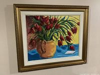 Framed oil painting showing a colorful floral still life with red flowers in a terracotta vase on a blue surface.