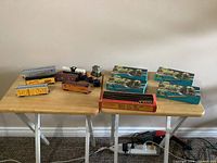 Photo of the entire lot showing the seven rail cars, plastic house, box of Tyco tracks and three Athearn boxes on a wooden table.