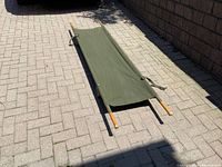Vintage military folding stretcher in open position on paved ground showing olive green canvas and wooden handles.
