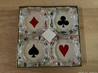 Set of four glass ashtrays each bearing a different poker suit symbol: diamond (red), club (black), spade (black), heart (red) arranged in a box insert.