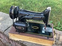 Side and top view of vintage Singer K99 sewing machine in black with gold decals, showing electric motor and power cord, mounted on wooden base with brown cover case seen underneath.