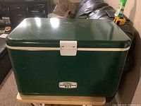 Full view of the vintage Thermos cooler showcasing its green exterior and white trim, placed on a small wooden table indoors.