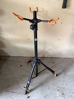 Black metal music stand with orange rubber padded arms and tripod base shown from the front, on a concrete floor against a beige wall.