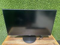 Front view of Asus VN279Q 27-inch monitor showing black screen and round stand base on wooden surface against green artificial turf background.
