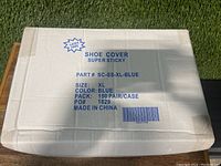 White cardboard box labeled 'Shoe Cover Super Sticky' with details including size XL, color blue, 150 pairs per case, made in China, latex free.