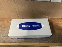 Single box of Uline facial tissue with blue logo on white box shown close-up on wood surface.