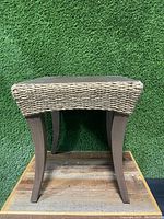 Side view of the wicker and wood patio end table showing the wicker woven top and wooden legs against a green grass-like background.