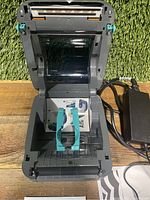 Printer interior with top cover open showing thermal print head and teal media guides, power adapter visible