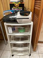 Full view of Rubbermaid plastic bin with four clear drawers on casters, Shark handheld vacuum on top, Dirt Devil handheld vacuum behind it, and boxed motion sensor security light.