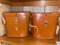 Two brown leather binocular cases placed side by side on a wooden shelf