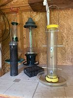 Three different bird feeders displayed on a floor against a wooden wall, showing variety in size and design.