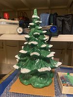 Green ceramic Christmas tree with white snow accents on branch tips, displayed on a shelf with background items.