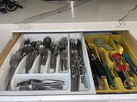 Drawer fully open showing sections with forks, spoons, knives, scissors, corkscrew and other utensils organized in a plastic tray.