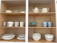 Three shelves with Corelle dinner and side plates, mugs and bowls on top two shelves. Ceramic cream and sugar set on top right shelf, assorted mugs including floral and solid colors, coffee mugs, and ashtray on the middle shelves.