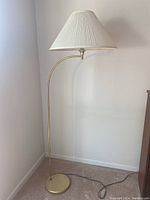 Full view of the brass floor lamp showing the curved arm and white pleated lampshade standing on carpeted floor by a wall corner.
