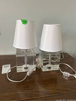 Two nightstand lamps with clear glass cube bases and white fabric shades photographed side by side on a wooden surface.