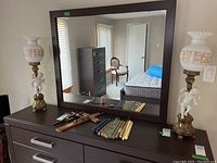 Pair of cherub lamps shown on dark wood dresser with mirror; both lamps have ceramic cherub figures on metal bases and white glass shades with gold detailing.