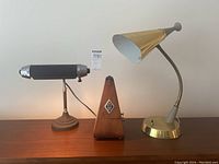 Shows both desk lamps and mechanical metronome standing on a wooden surface. Larger lamp on right with brass cone-shaped perforated shade, smaller lamp on left with black cylindrical shade and silver ends, metronome in center.