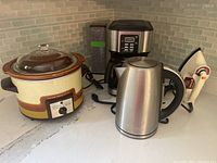 Overview showing all appliances arranged on countertop: Canadian General Electric crock pot, Sunbeam coffee maker, Cuisinart electric kettle, Black and Decker iron, and straw tin.