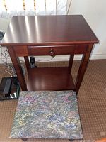 Full view of wood side table with drawer and lower shelf and upholstered wood footstool in front.