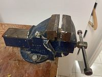 View of the whole metal vise mounted on wooden worktop base showing the threaded clamp handle and textured gripping jaws.