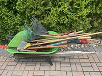 Overall view of green metal wheelbarrow loaded with various garden tools including rakes, shovels, broom, and pitchfork, showing outdoor wear