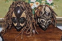 Photo showing both African masks side by side on wooden surface with floral wallpaper background. Left mask has braided hair, right mask has bead hair.