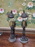 Pair of antique metal table lamps viewed front on a table against floral background, no shades, cords coiled at base