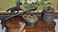 All three items on wooden surface with floral background: black iron weather vane horse on arrow, small round tin kettle, and large cylindrical tin kettle.