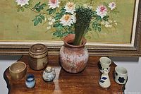 Photo showing all 8 pottery pieces arranged on wooden surface beneath a floral framed painting, illustrating variety in size, shape, and glaze style.