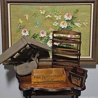 Photo showing all four wooden items placed on a surface with a framed floral artwork in the background. Items visible include the small three-tier wooden shelf, a wooden crate with vintage label, the house-shaped wooden box, and the small wicker-sided crate.