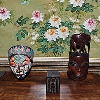 Three-item lot on wooden table against floral wallpaper: hand painted tribal mask, vintage carved wooden box, and carved wooden face with elephant.