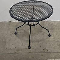 Side view of metal outdoor patio table showing four curved legs, mesh top, and concrete floor setting.