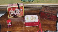 Four vintage tin boxes on a wooden surface: Union Leader Cut Plug tobacco tin, Petticoat Junction lunch box, Little Wonder Story tin, and a small red tin container.